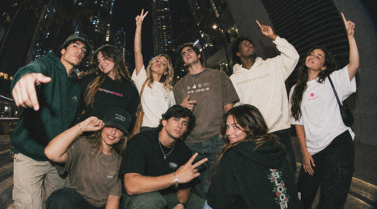 A group of eight Christian young adults posing together in a nighttime urban setting with city skyscrapers visible in the background. They are wearing casual streetwear, including hoodies, t-shirts, and sweatshirts in various colors like black, white, brown, and green.